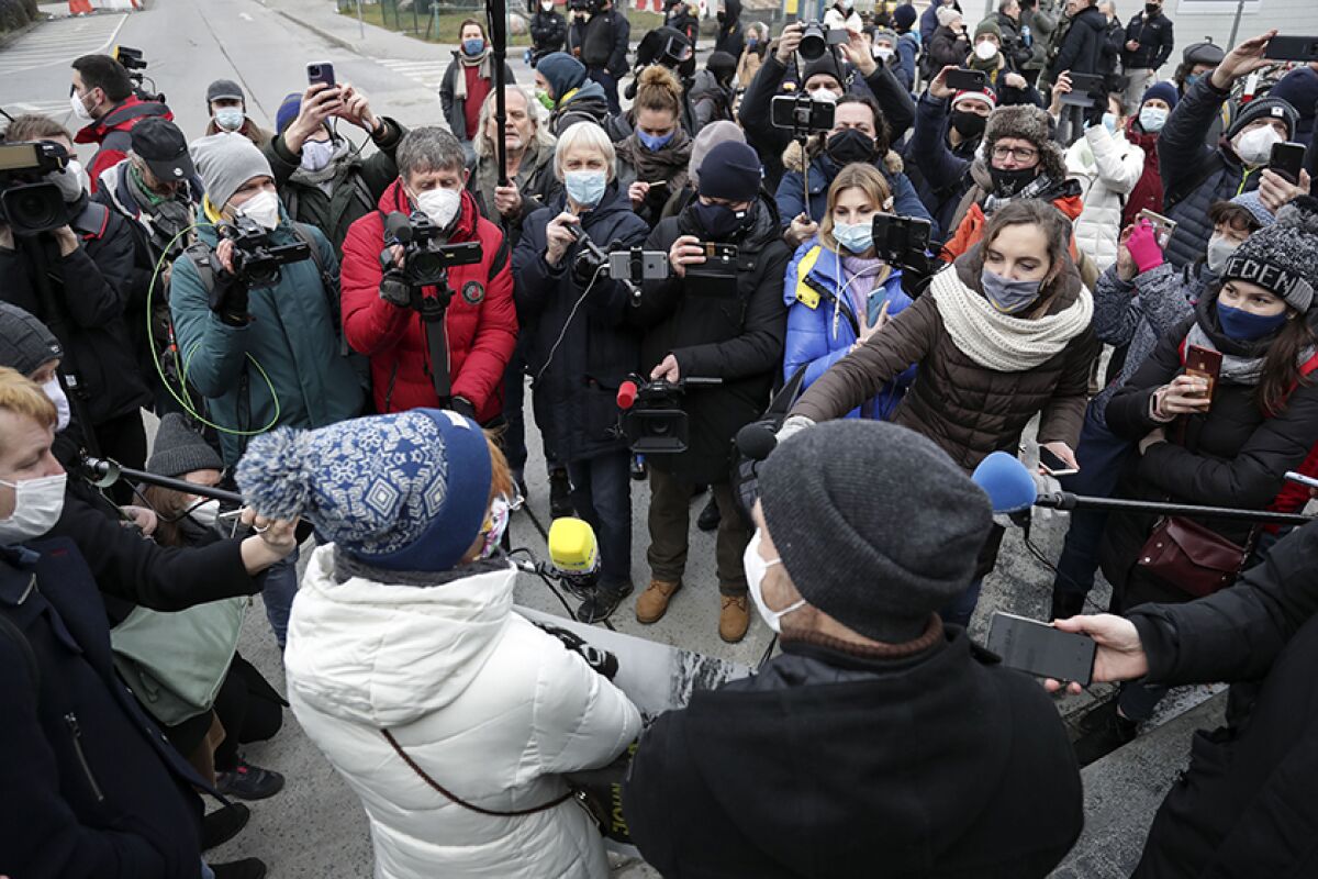 People in coats and masks speak into microphones amid a crowd.