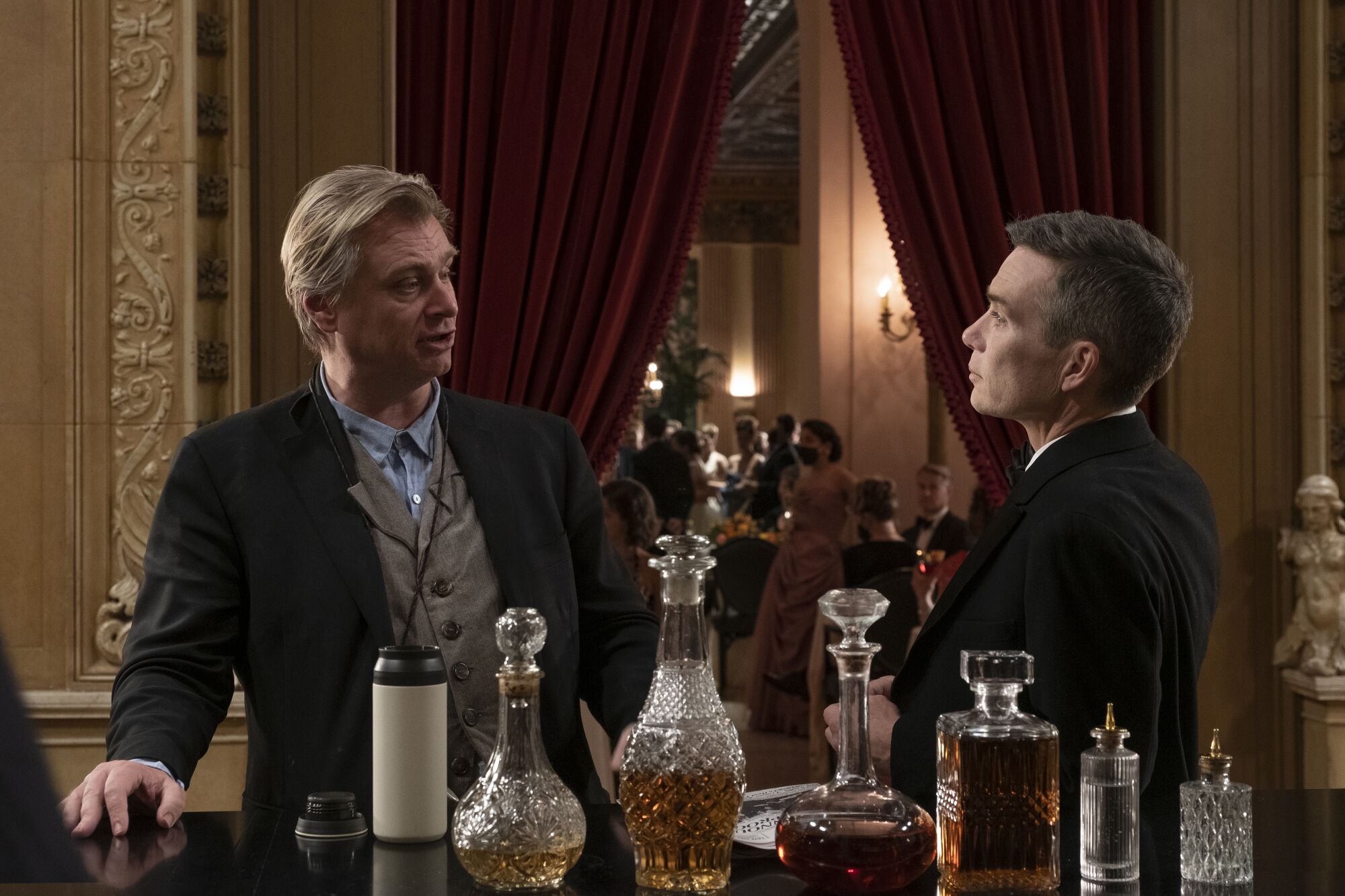 Bir film setinde takım elbiseli iki adam, arkalarında kırmızı perdeler ve önlerinde alkol şişeleri olan bir odada.