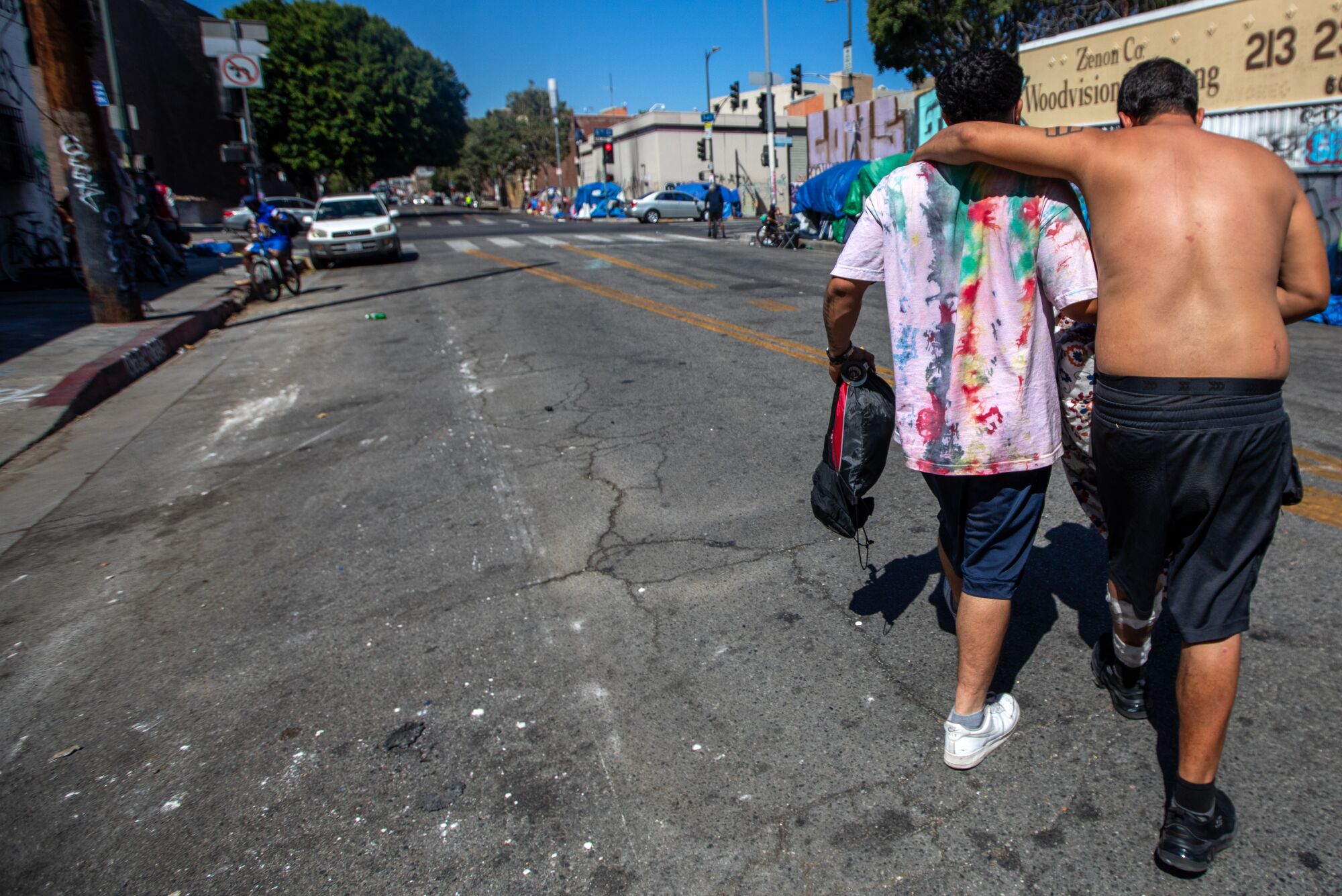 A shirtless man leans on another man for support as they cross a street.