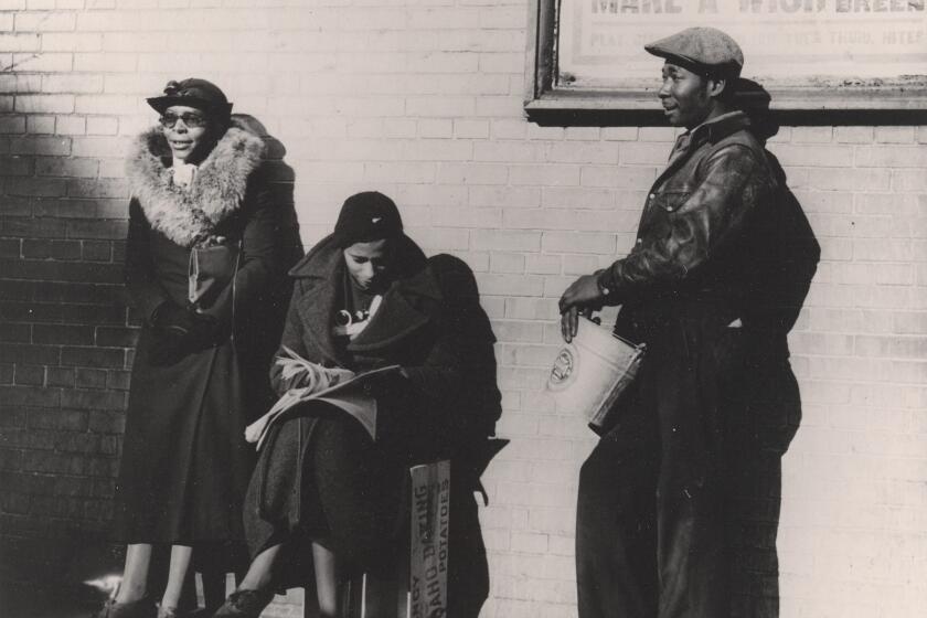 A vintage black and white image show two Black women and one Black man in the sunshine before a brick wall