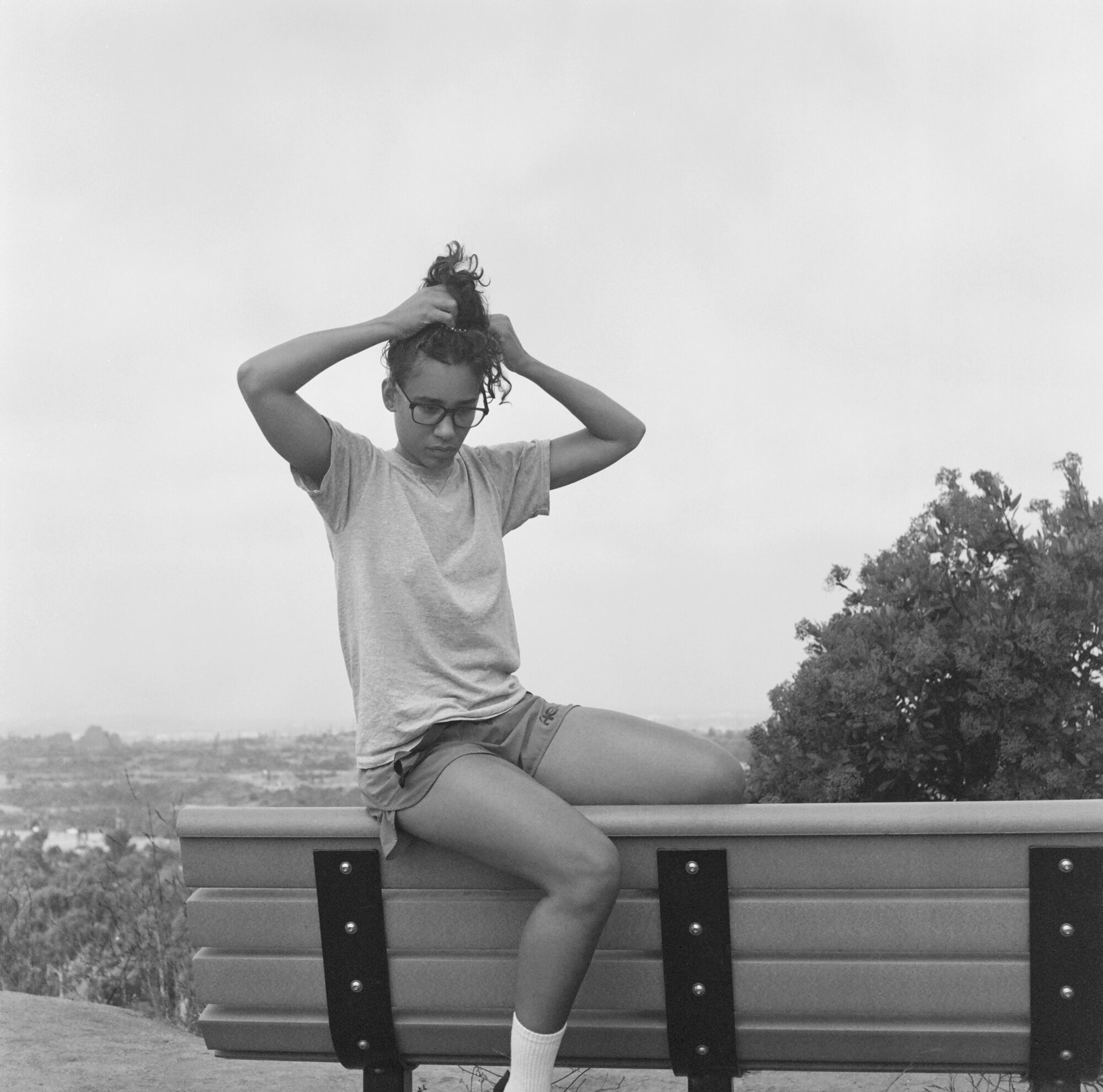 A black and white image of Lacey Lennon sitting on the edge of a bench tying her hair up at Kenneth Hahn park