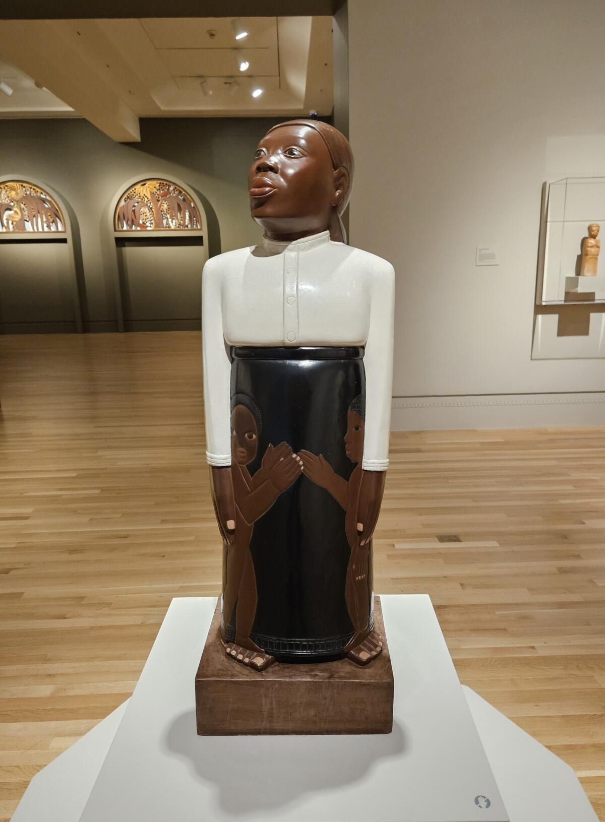 A wood, totem-like sculpture of a mom in a white blouse and black skirt sheltering two kids by each leg.