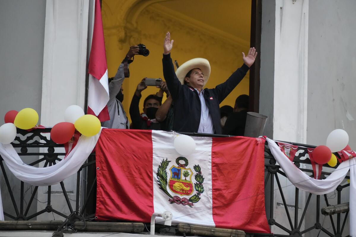 A man in a wide-brimmed hat stands with his arms up and outstretched by a railing draped with a red and white flag 