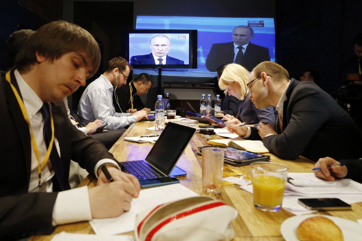Reporters sitting at a table take notes while listening to Vladimir Putin
