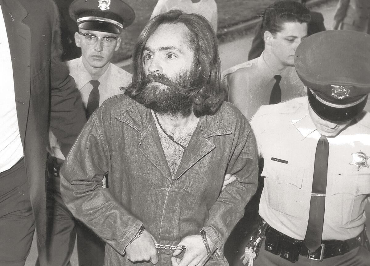 Police escort a handcuffed man with long hair and a beard
