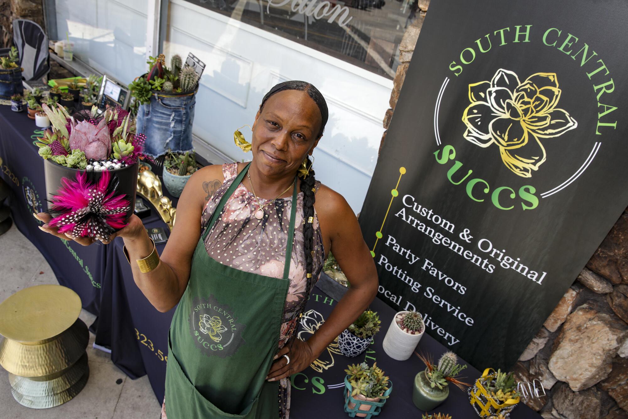 La Nola Smith holds a succulent arrangement while standing in front of her sales table that holds more arrangements.