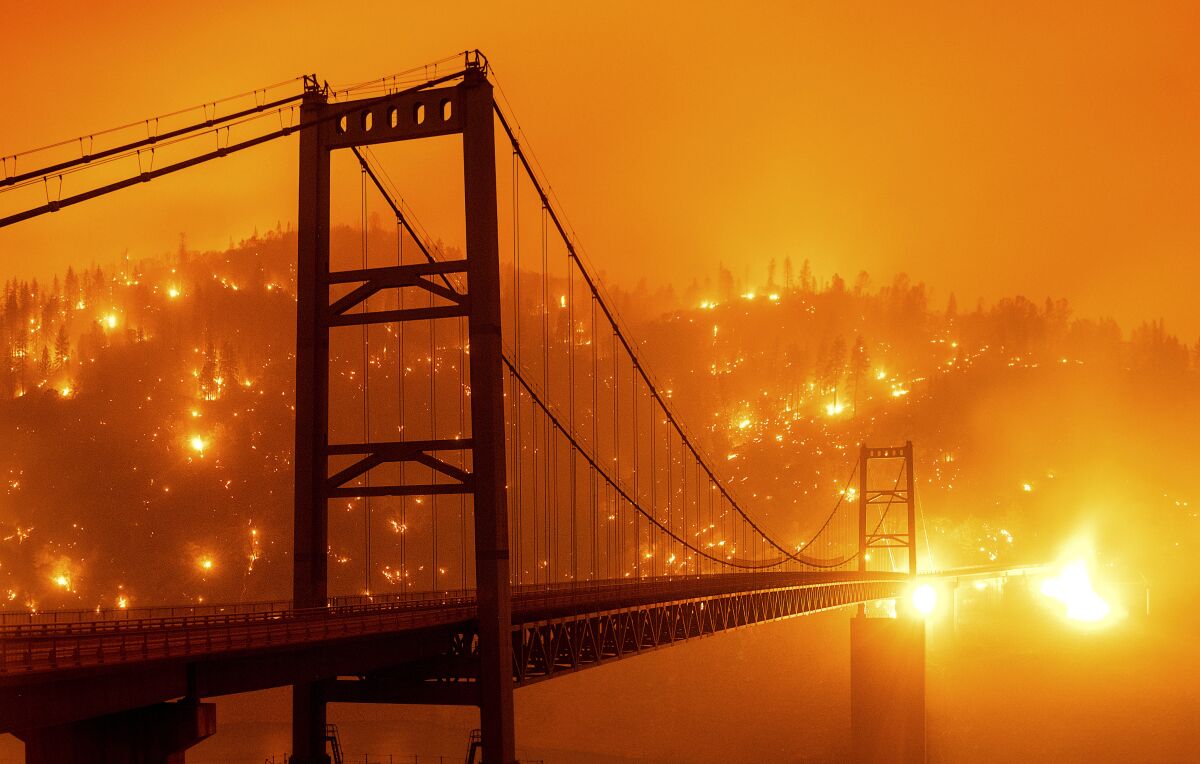 Flames burn across a hillside behind a suspension bridge, with an orange glow in the sky