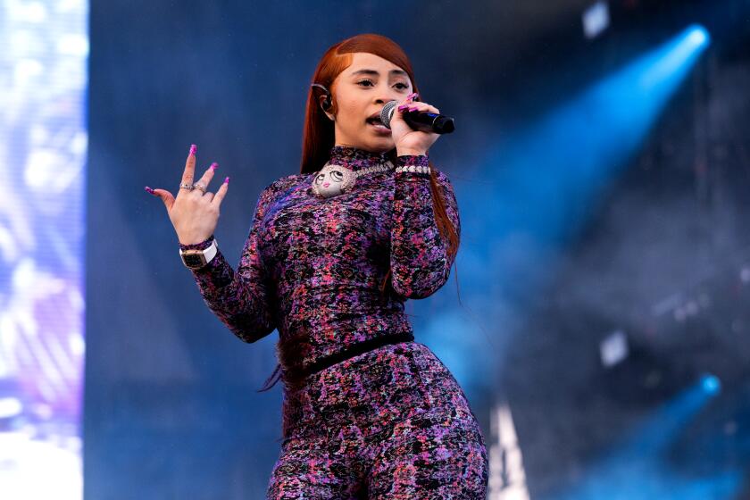 Ice Spice in a dark patterned jumpsuit holding a microphone with her left hand and holding up her right hand