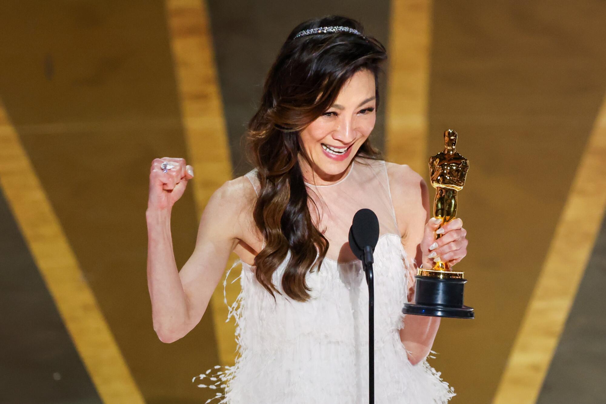 A woman in a white dress with long dark-brown hair, smiles and gestures as she accepts an award