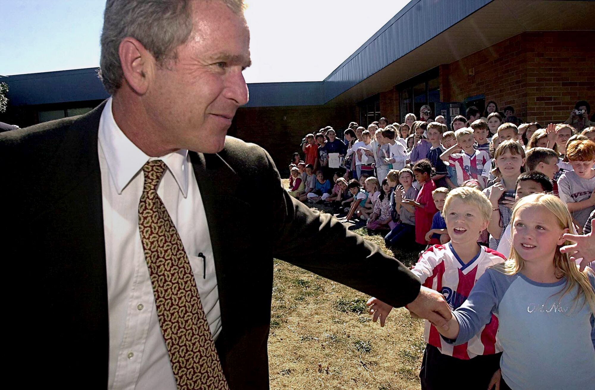 George W. Bush walking away from a crowd of children outdoors as some reach out to touch his hand.