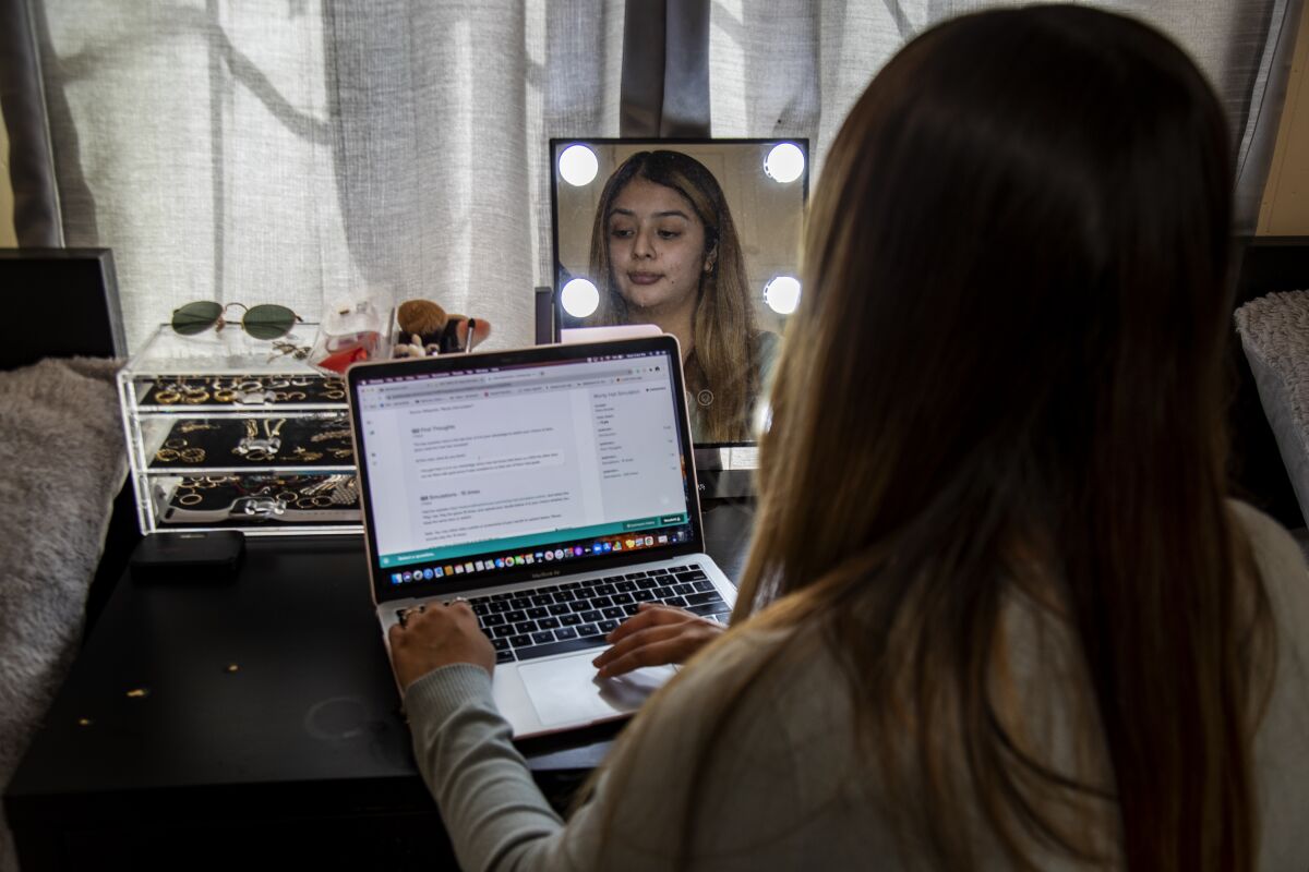 A student works on her laptop at home, her face reflected in a mirror on her desk