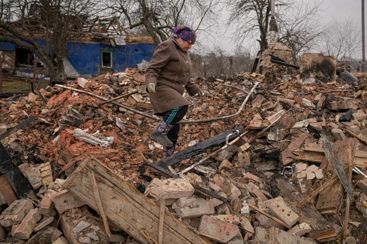 A woman in a brown jacket and gloves looks down as she navigates a pile of rubble