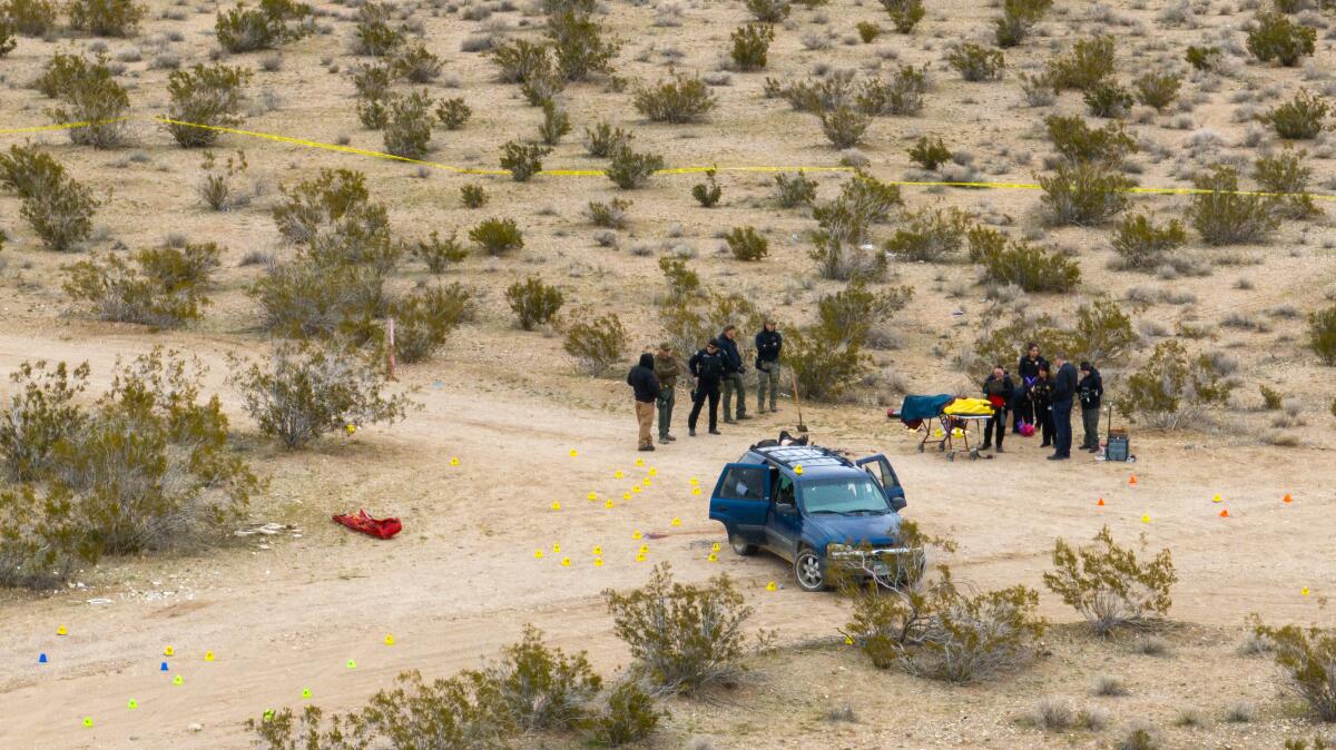 Several officials stand near a van and crime scene tape in the desert, the ground scattered with tags in yellow, red and blue
