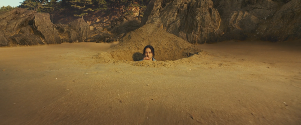 A woman stands in a hole at the shore with sand up to her neck in "Decision to Leave."