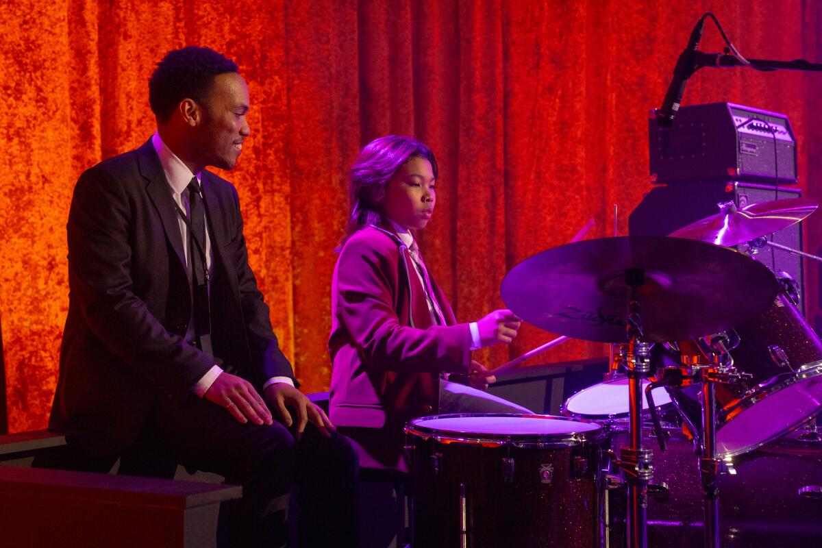 A father looks proudly at his young son, both sitting at a drum kit