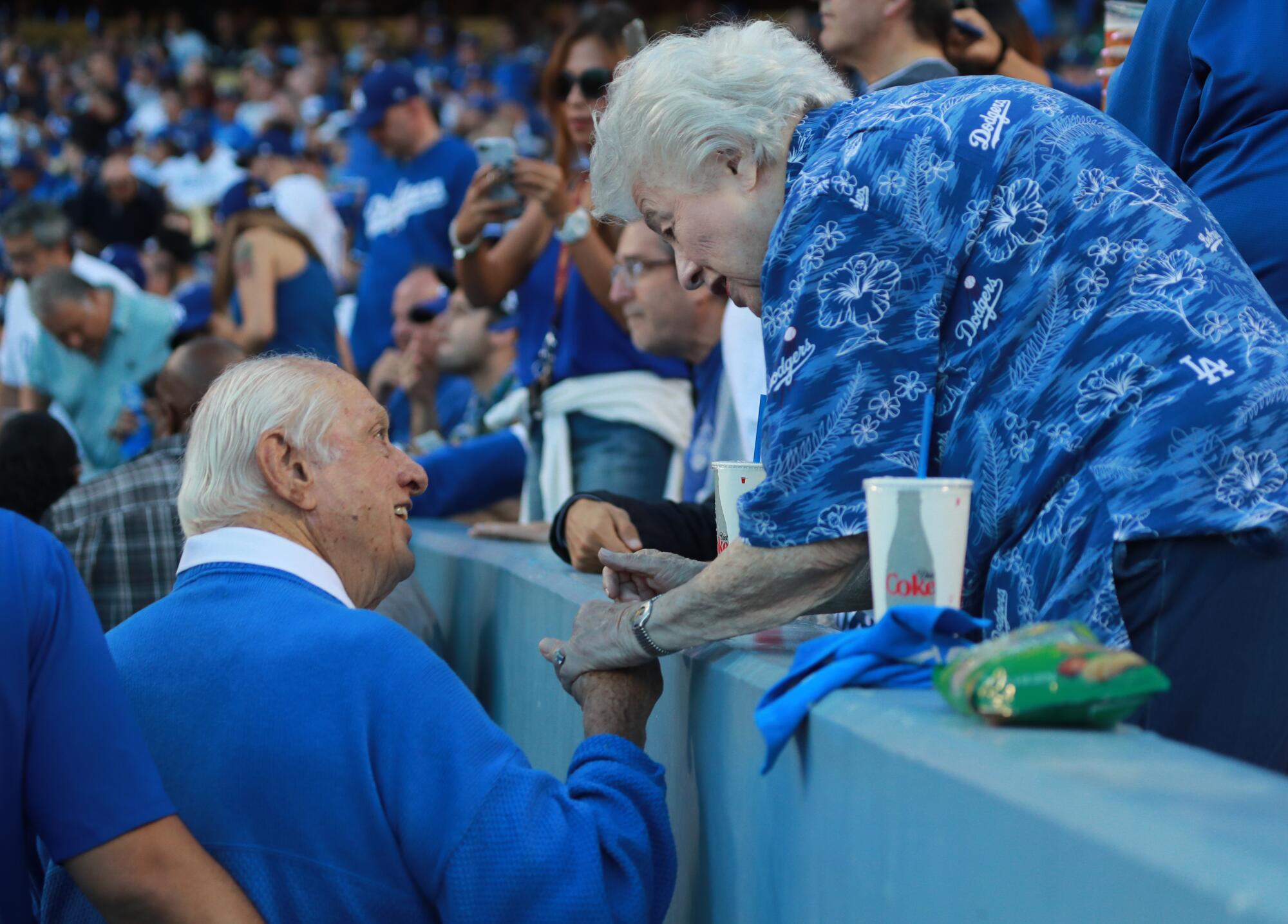 Tommy Lasorda holds Rosalind Wyman's hand and they talk in stadium stands.