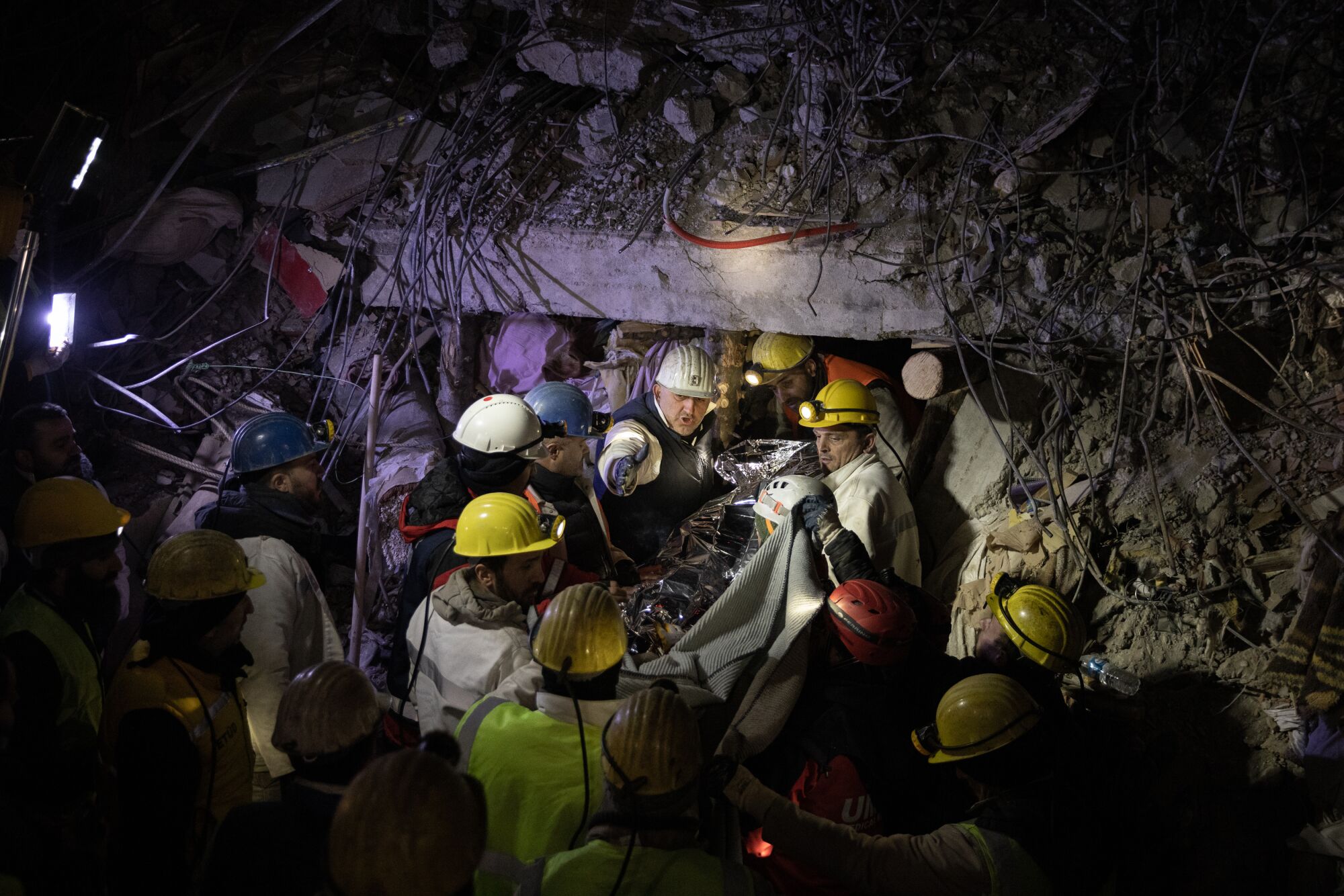 Several people in hard hats help carry out a person on a stretch from rubble at night