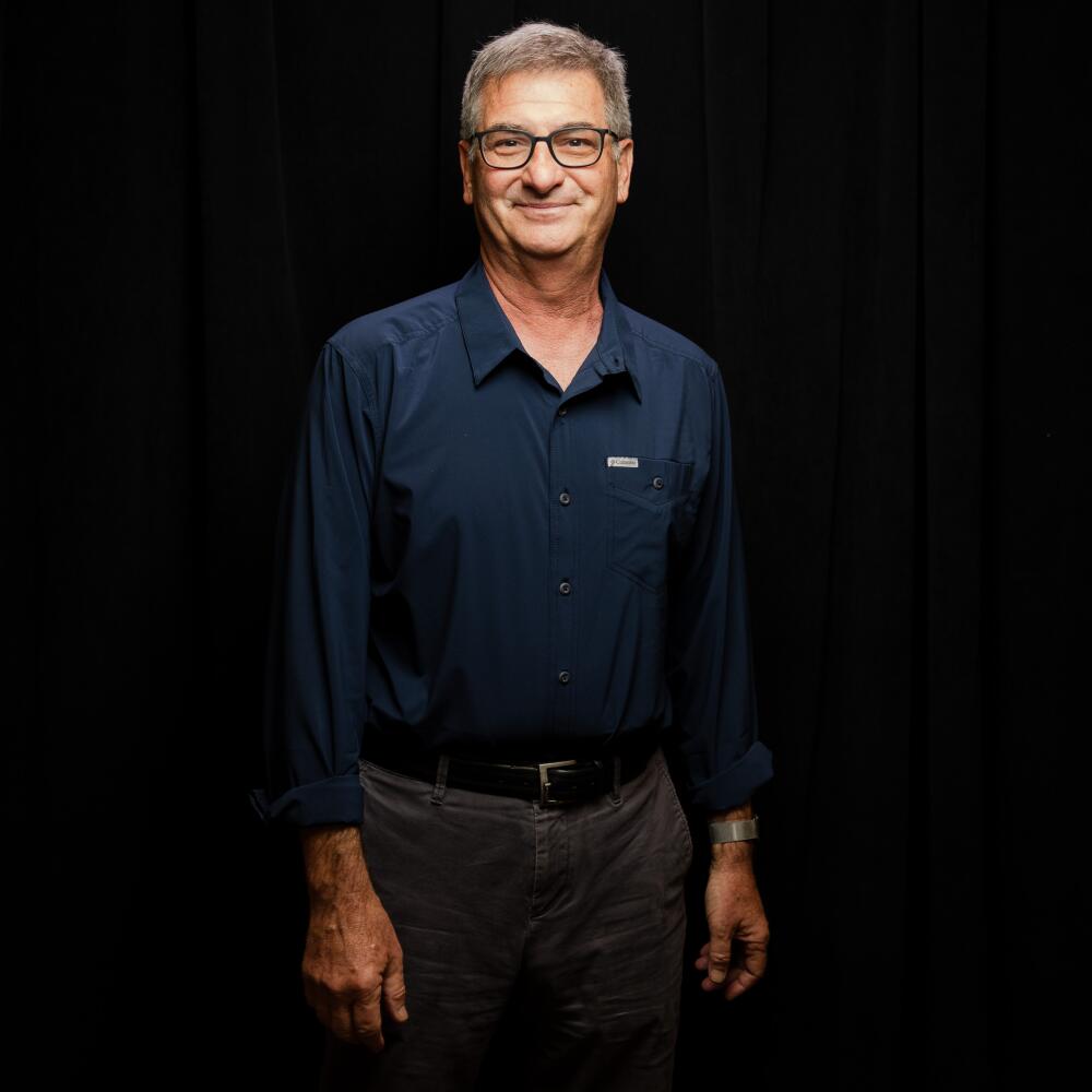 A smiling man with glasses, wearing a long-sleeved blue shirt, against a dark backdrop