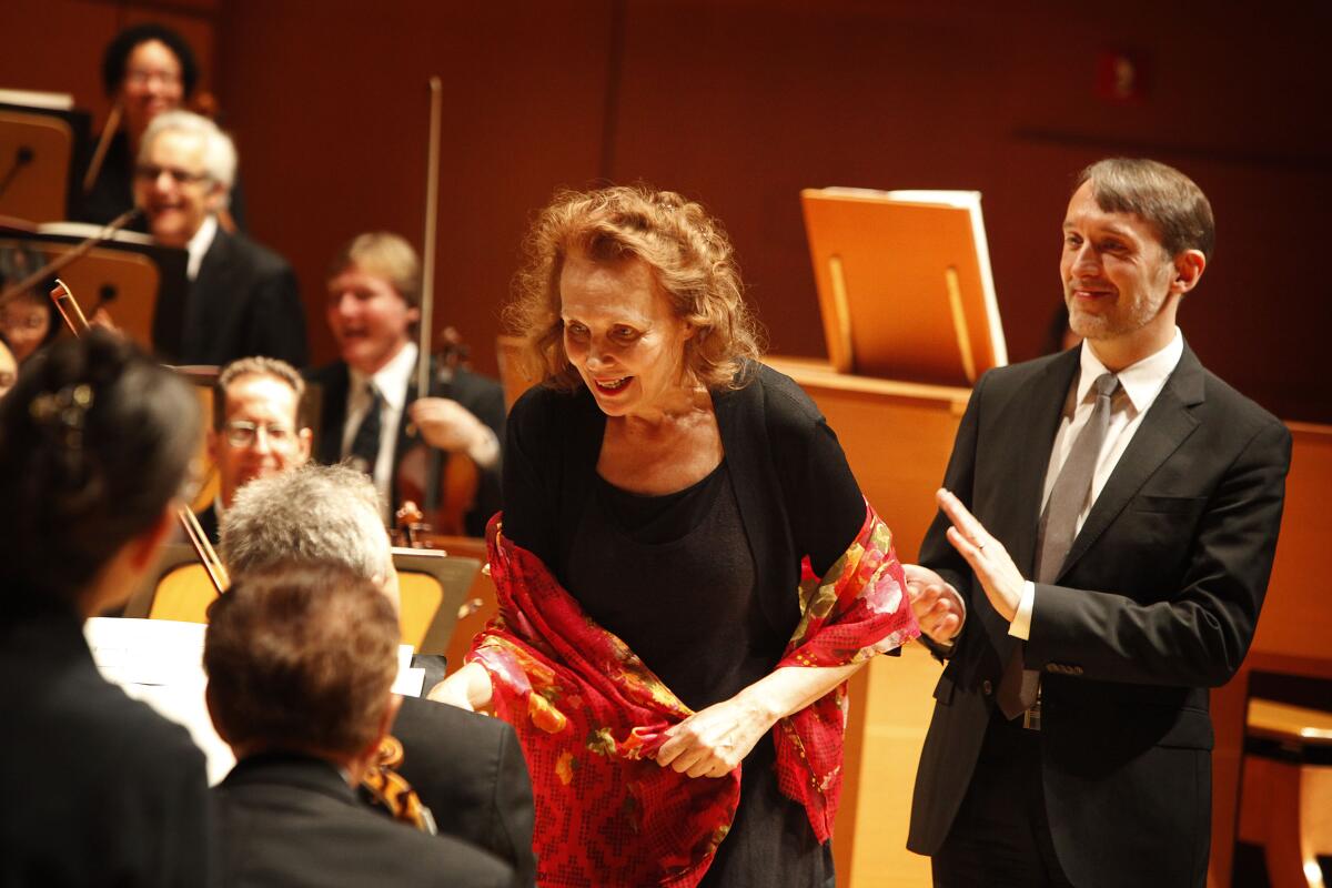 A man in a suit claps for a composer as seated musicians congratulate her