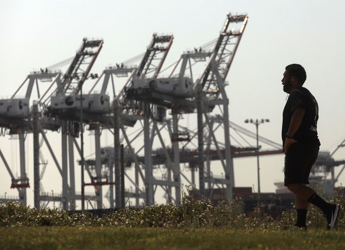 A man walks along the Wilmington Waterfront Park against a backdrop of cranes at the Port of Los Angeles.