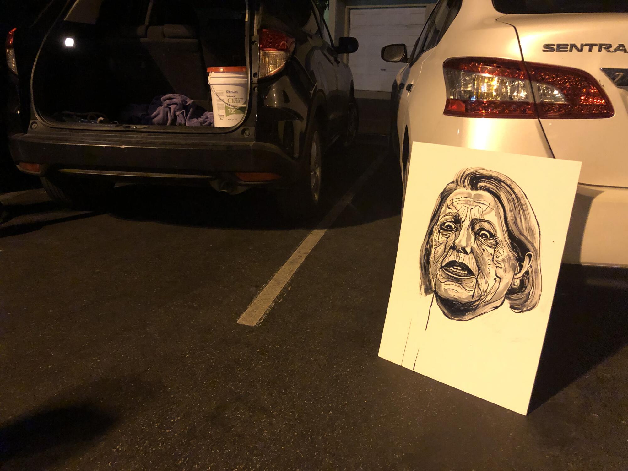A white poster featuring a caricature of Ginni Thomas' face rests against a car bumper in a parking lot.