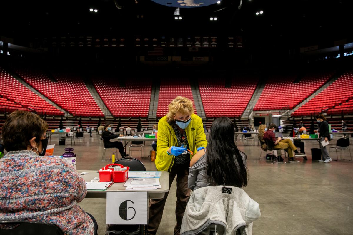 A nurse dressed in a yellow jack gives a vaccine shot to a woman in an  arena.