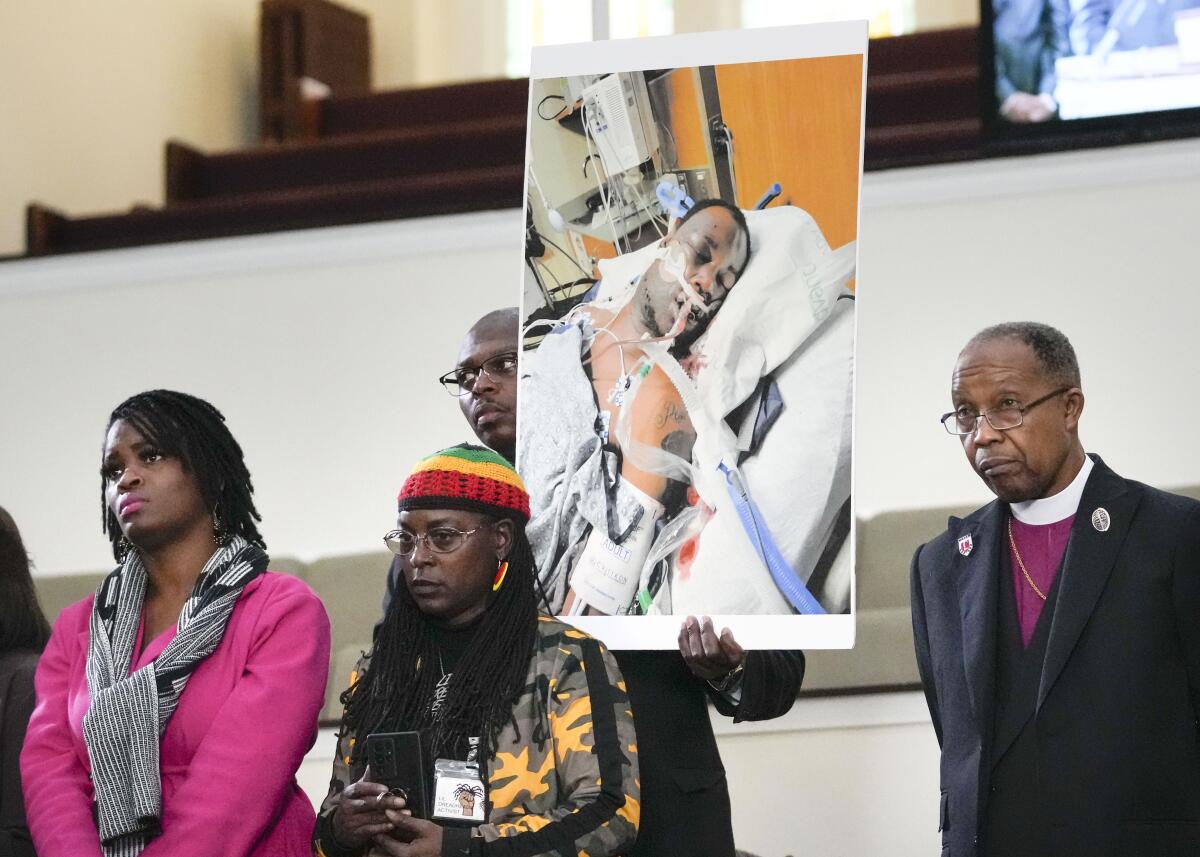 Five people, including a pastor, stand together, one holding an image of a Black man lying in a hospital bed.