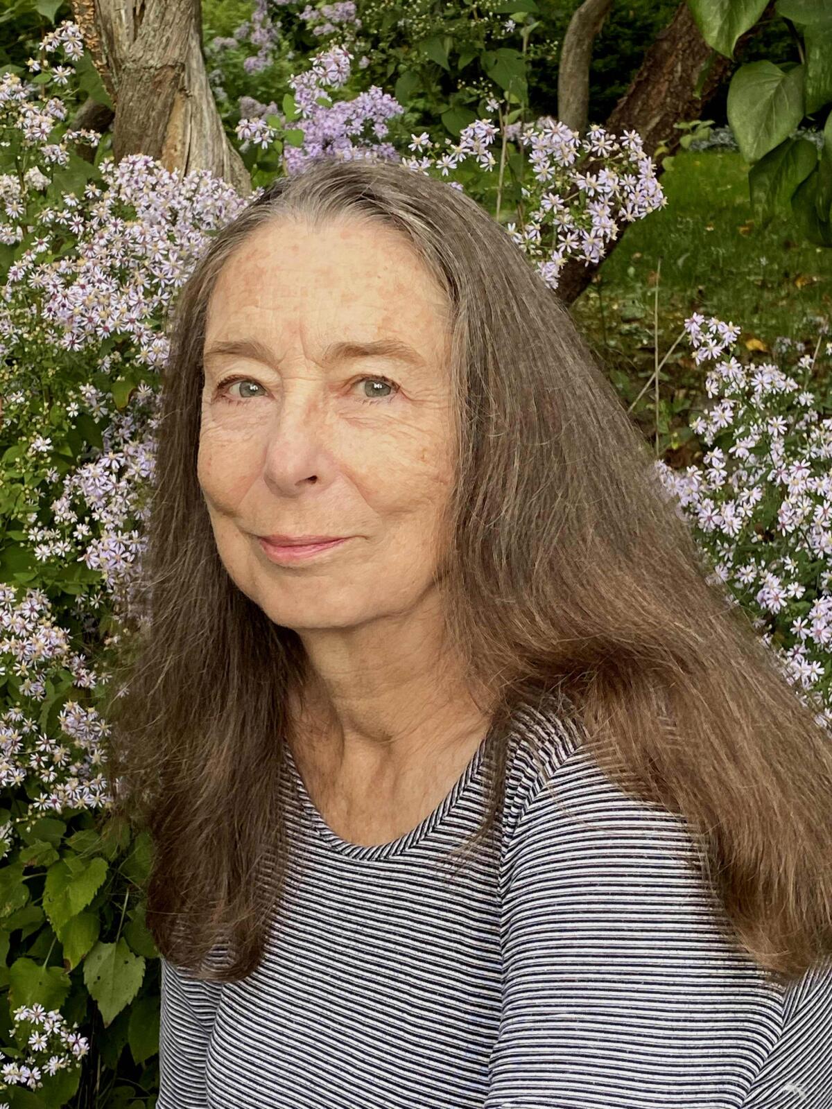 An older woman in a striped sweater standing in front of small pink flowers.