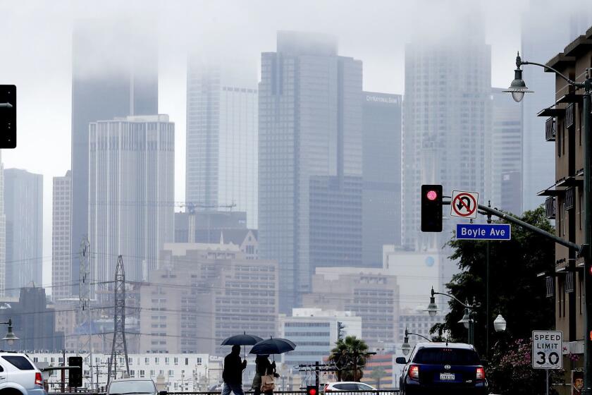 LOS ANGELES, CALIF. - MAR. 6, 2019. Pedestrians cross First Street in Boyle Heights as rain clouds partially obscure the downtown L.A. skyline on Wednesday, Mar. 6, 2019. (Luis Sinco/Los Angeles Times)