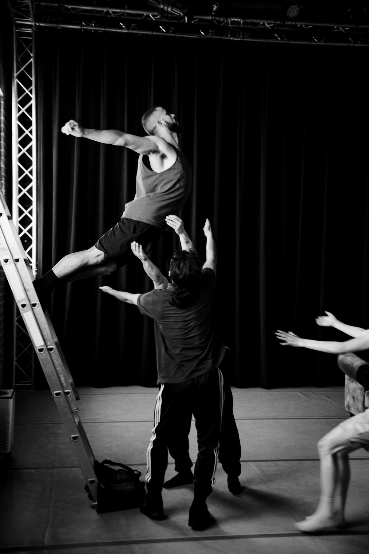 A dancer jumps off a ladder into the arms of other dancers on the ground.