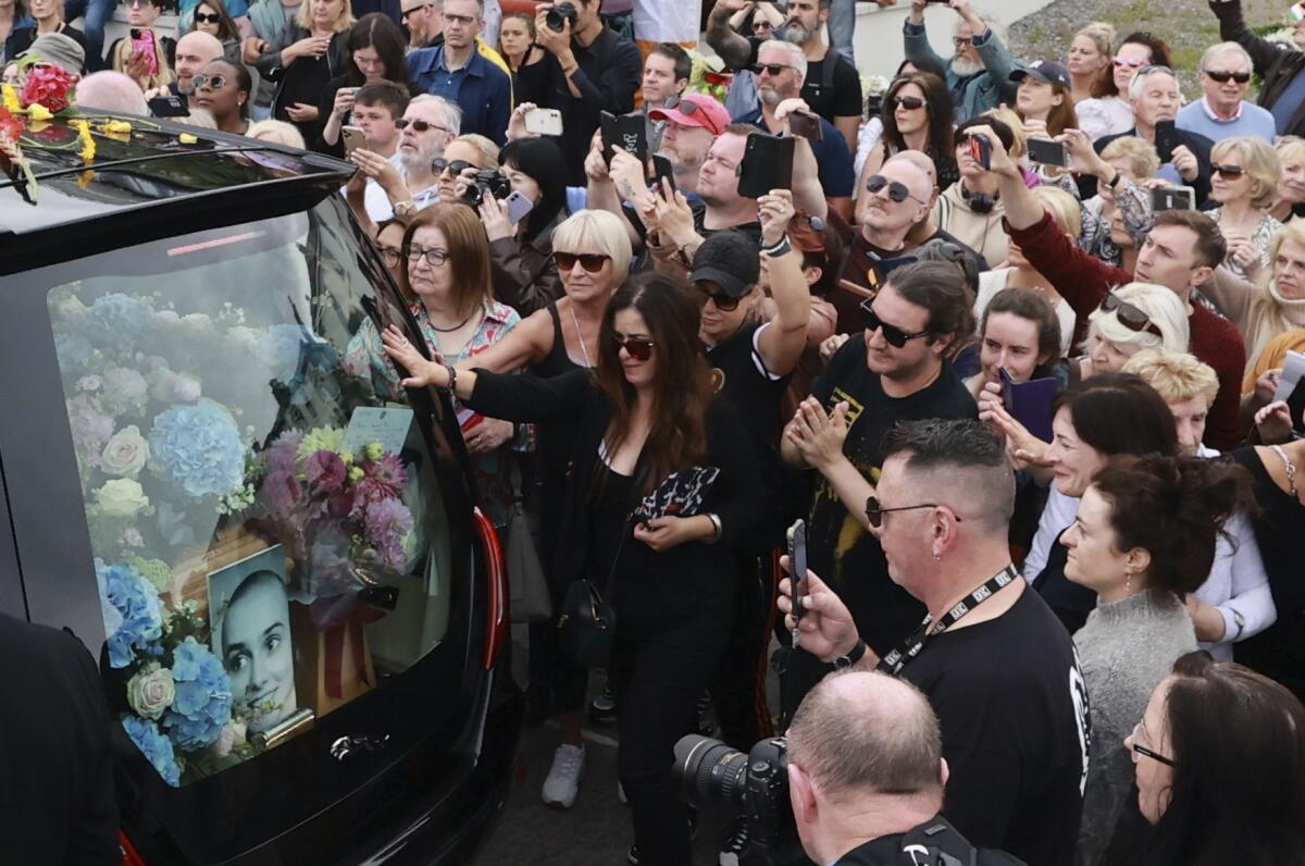 A crowd of mourners surrounds a hearse carrying a casket, flowers and a photo of Sinéad O'Connor