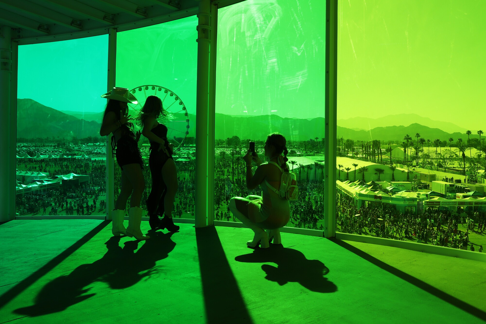 A woman takes pictures of two other women in a green-glass enclosed building overlooking the crowded grounds below.