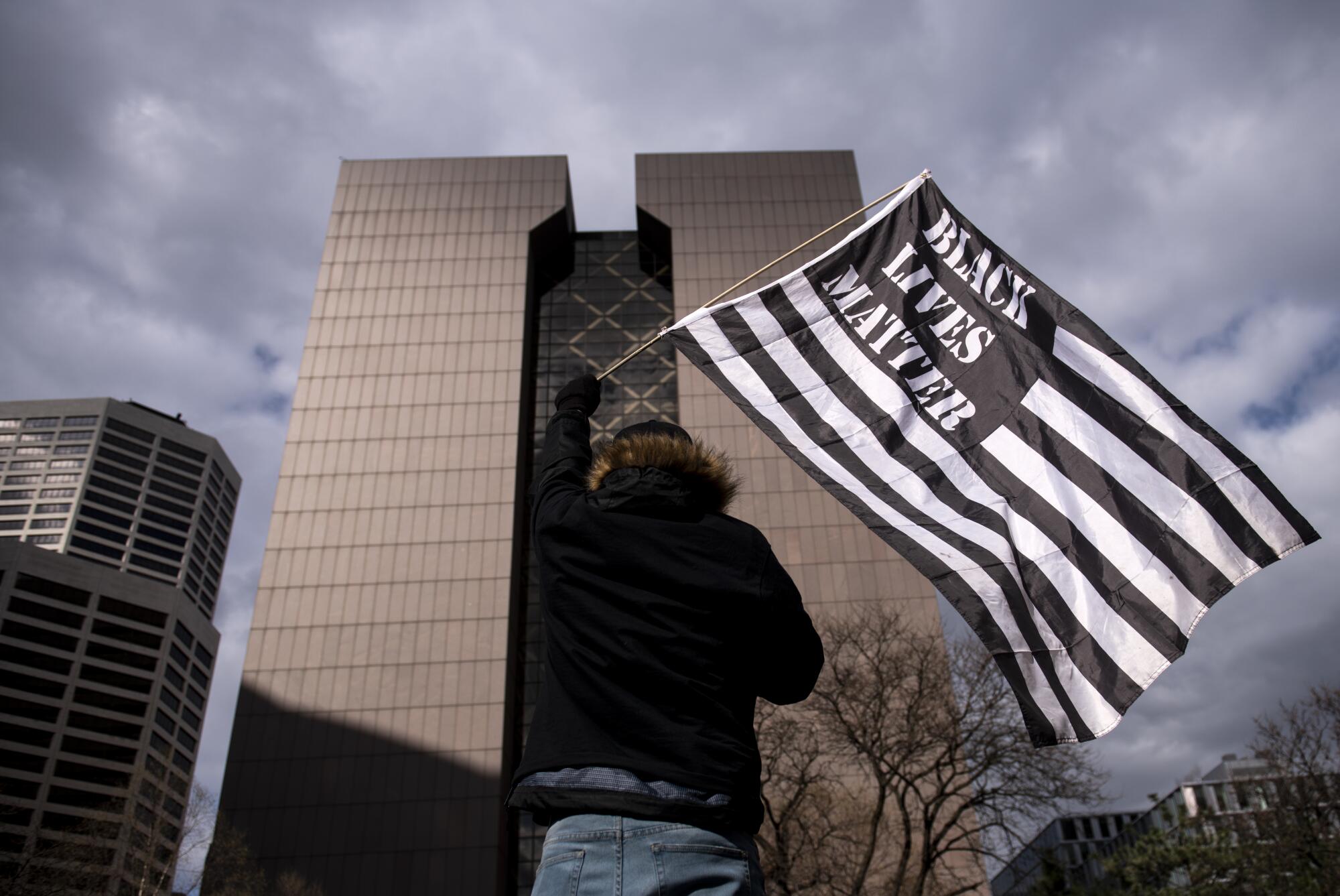 A person hoists a black-and-white striped flag with the words Black Lives Matter in front of tall buildings 