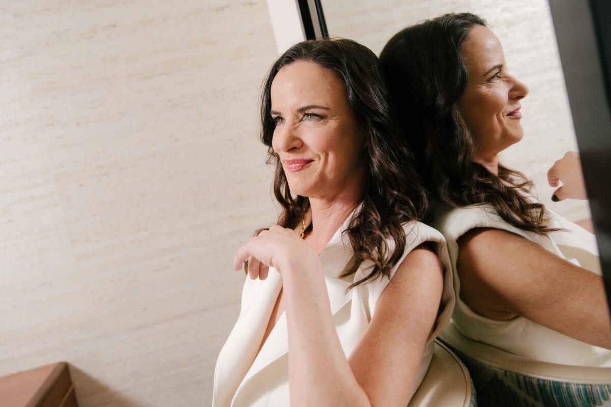 Juliette Lewis in a white sleeveless top, sits and leans against a mirror.