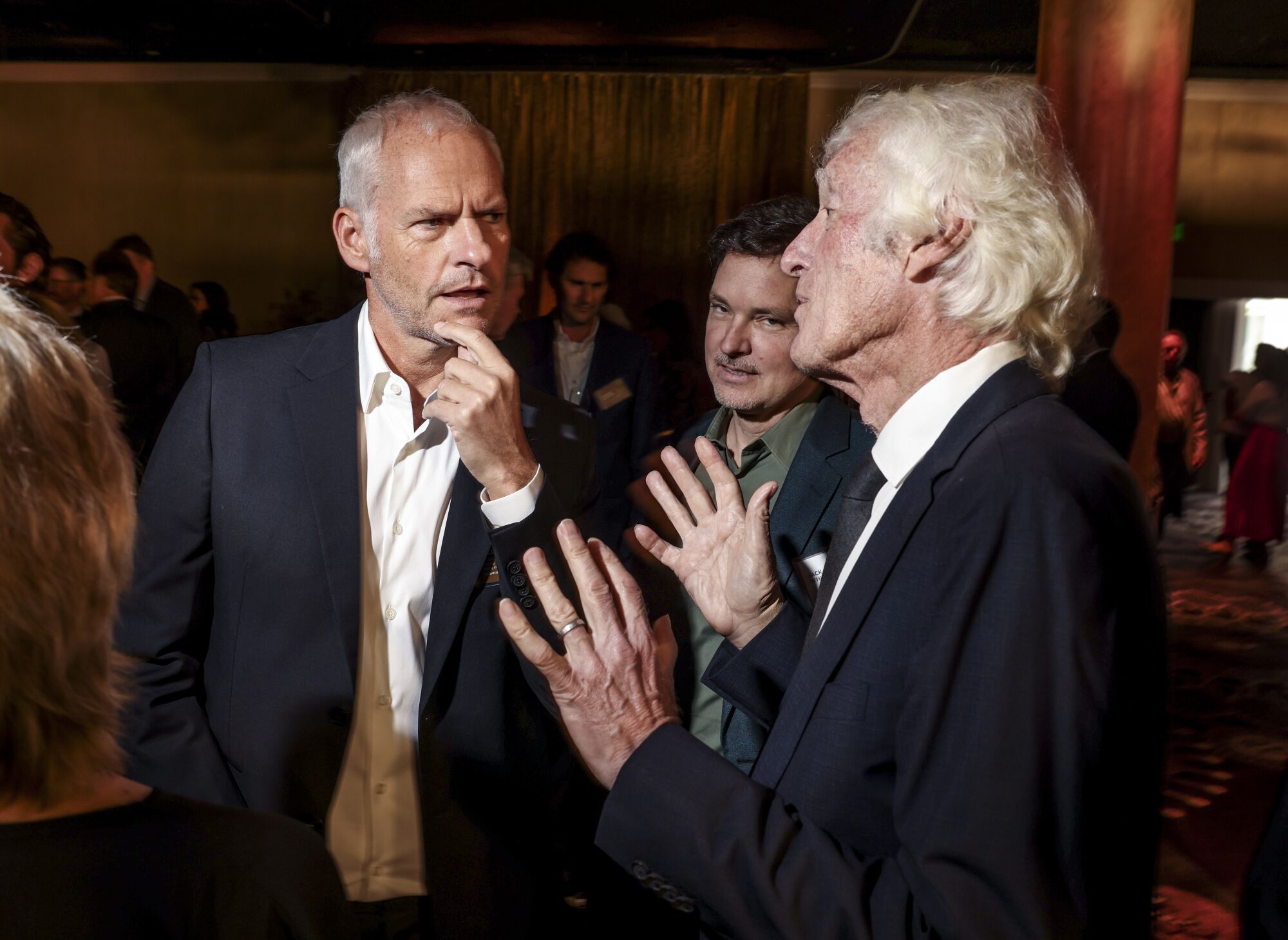 Two gray-haired men talk as another man looks on.