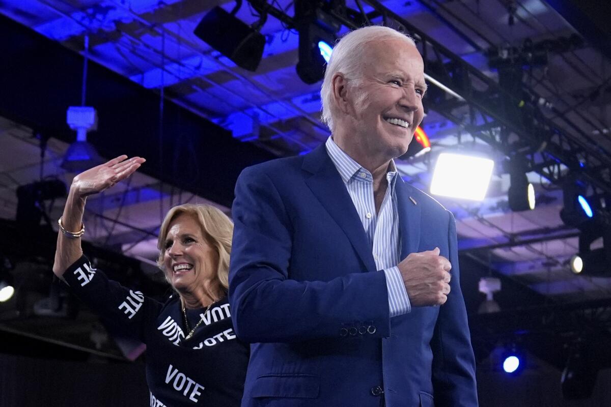 President Biden smiles and First Lady Jill Biden waves in an outfit reading "VOTE" as they exit a stage under cobalt lights