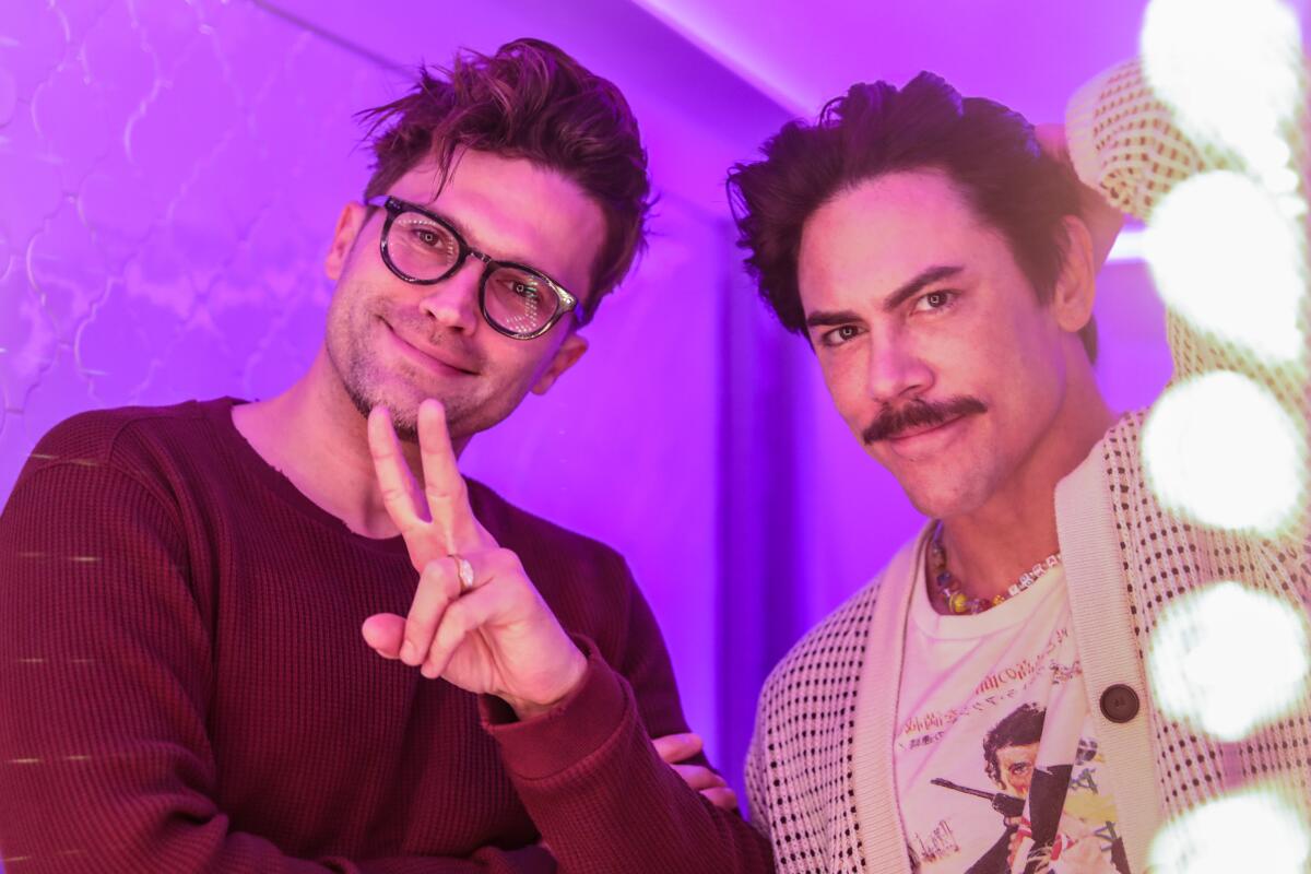 A man in glasses flashes a peace sign next to a man with a mustache and sweater as they pose in front of a mirror.