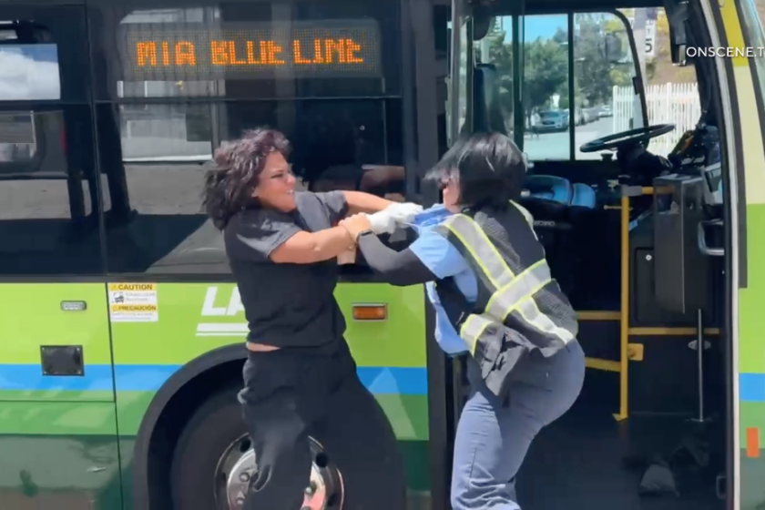 Two women, one in a reflective vest, grasp arms in a fight outside a green bus.