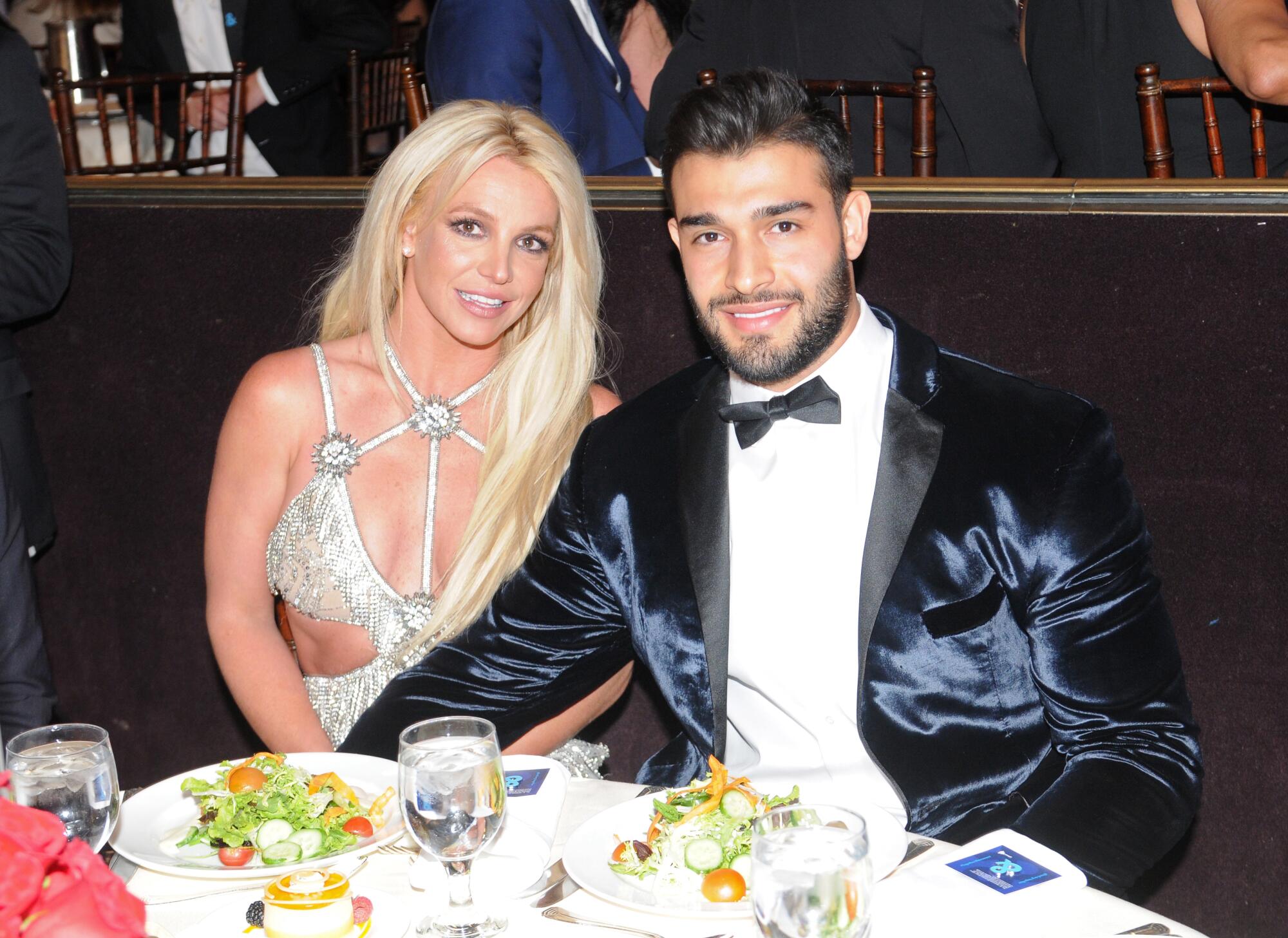 Britney Spears, wearing a silver dress, is sitting at a dinner table with Sam Asghari in a blue velvet suit