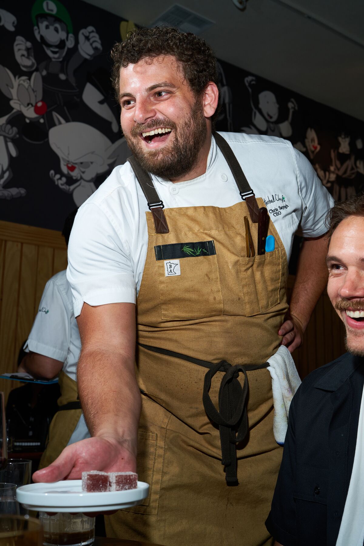 A smiling, bearded chef wearing an apron delivers a plate to a table.