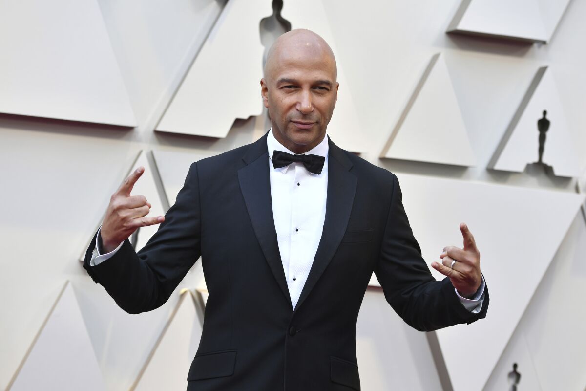 A bald man in a black tuxedo and bow tie makes rock-on symbols with his hands