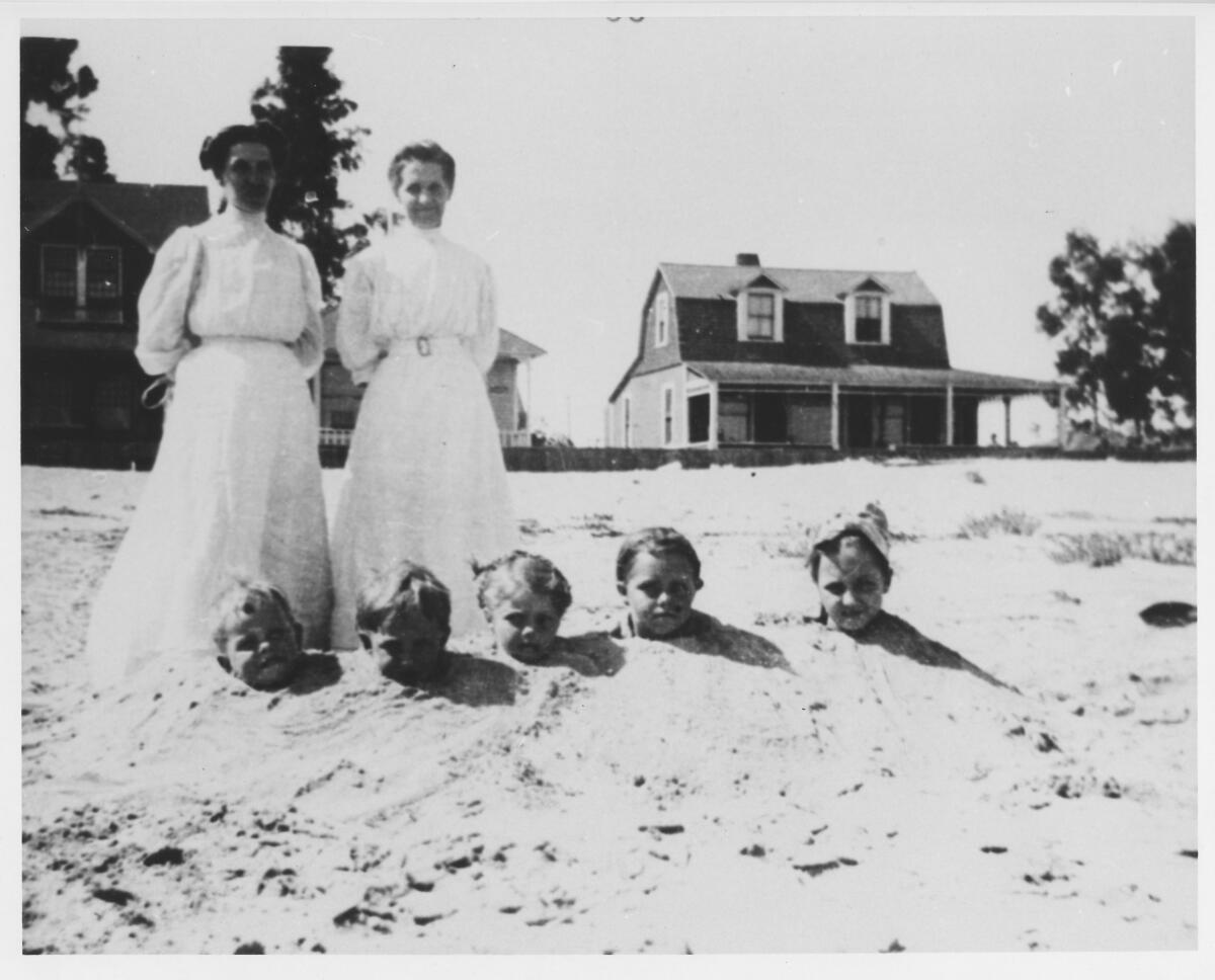A vintage photo shows two women in old-fashioned dresses standing on a beach with children buried up to their necks in sand.