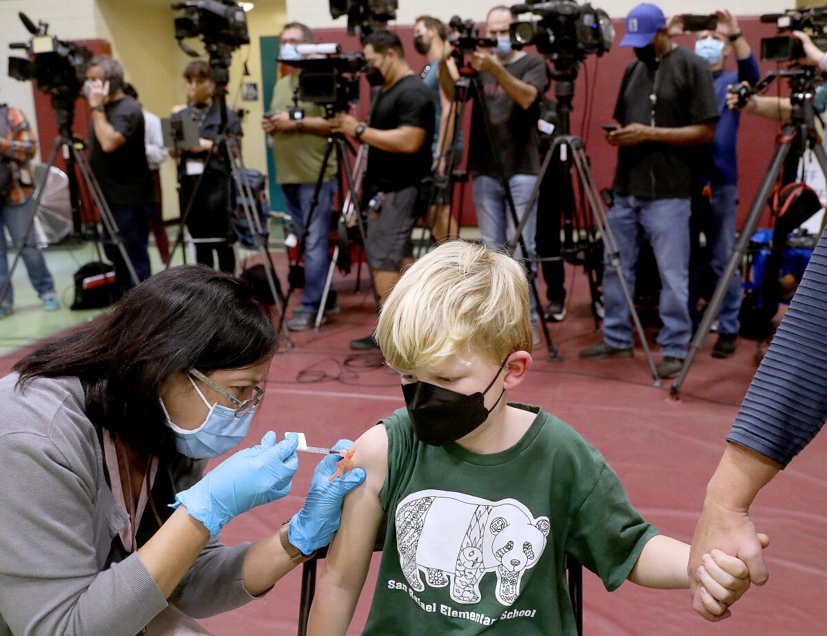 A little boy in a T-shirt with a panda bear receives a vaccination from a woman in a mask and gloves.