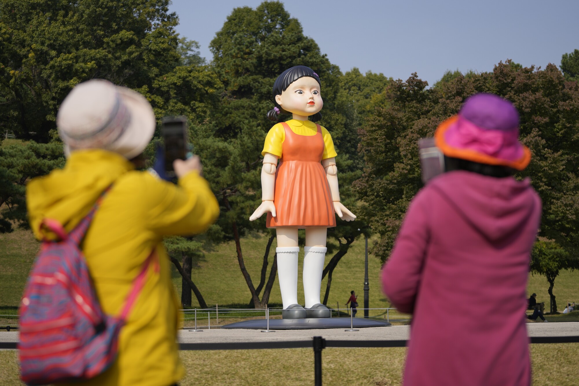 Two people take photos of a giant doll outdoors.