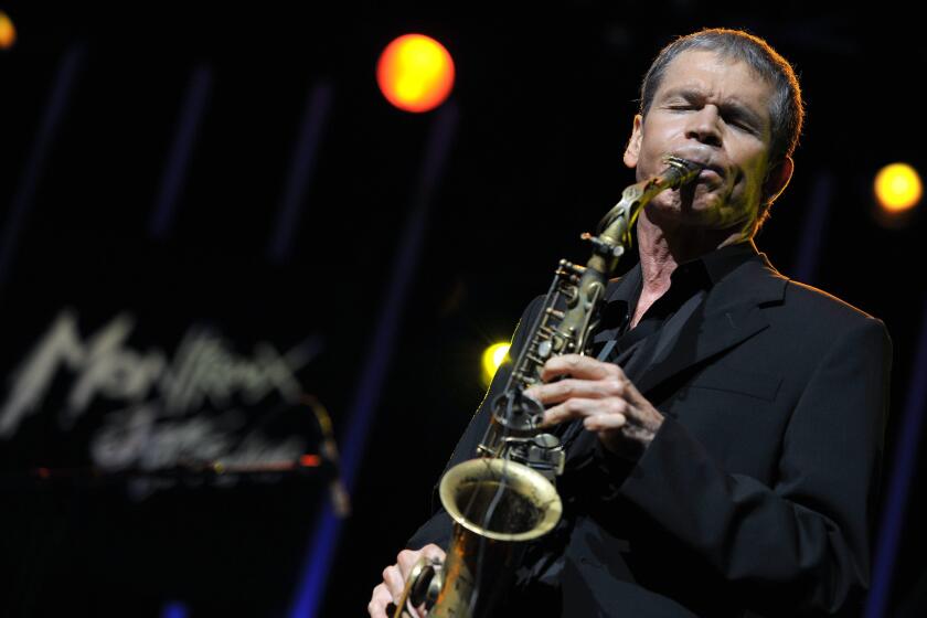 A man in a black blazer holding and playing a saxophone on a dark stage