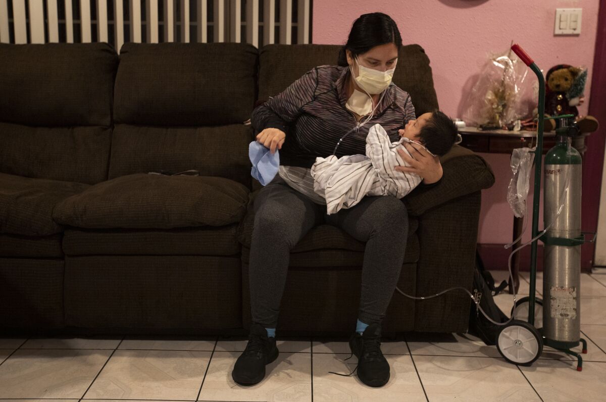 A woman in a mask sits on a couch with her newborn son, with an oxygen tank by her side