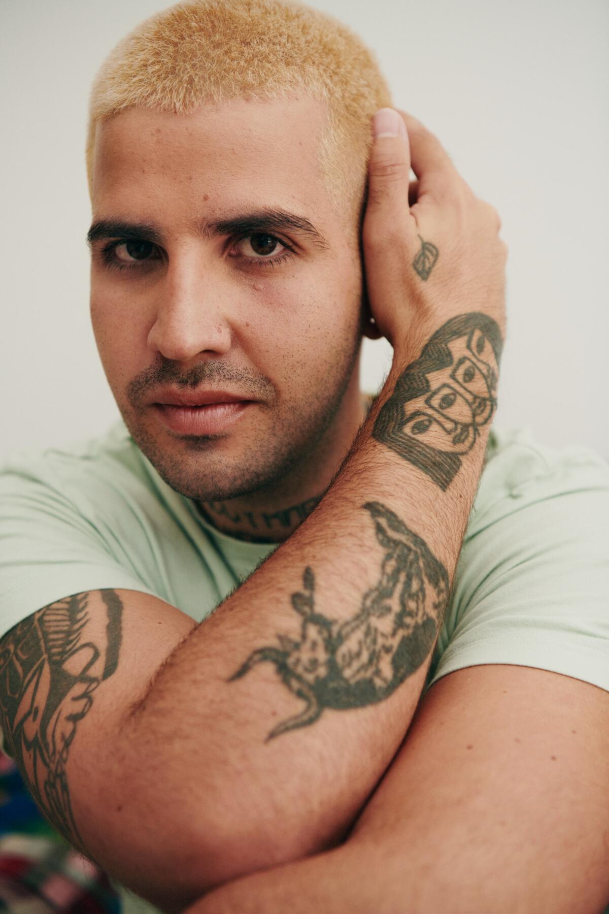A person with close-cropped fair hair touches the left side of their head with their tattooed right arm.