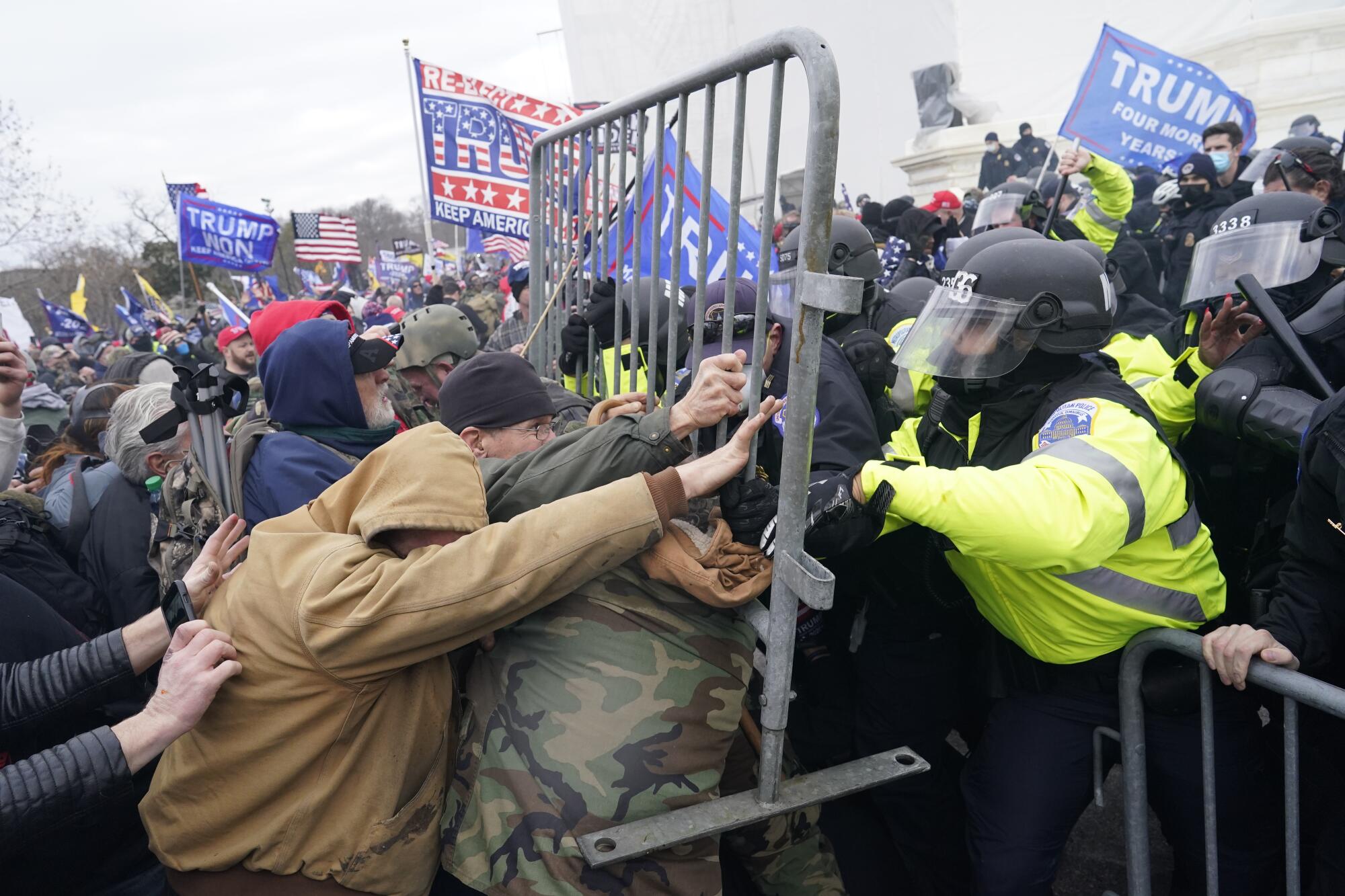 A crowd of rioters with Trump flags presses into a police metal barricade, with officers pushing back.