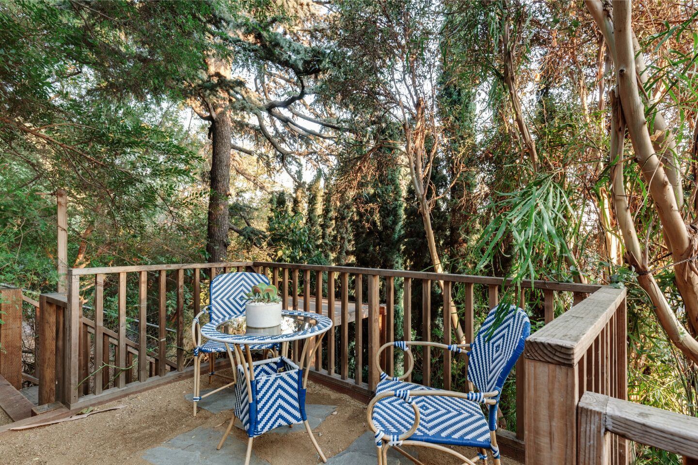 The dining patio with chairs and a cafe table, a wooden railing and trees behind it.