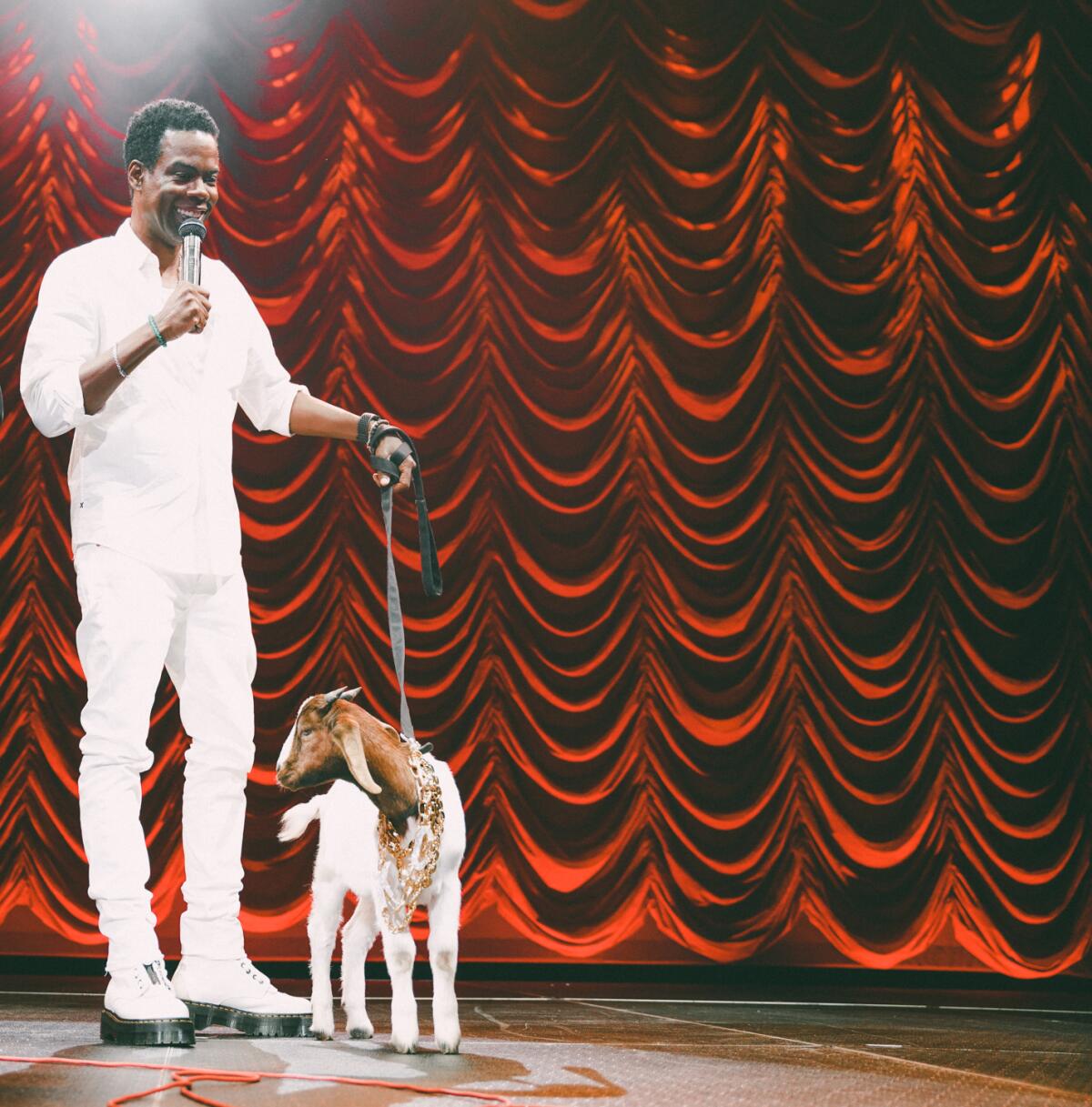 A Black man in a white outfit holds a microphone in one hand and a goat on a leash in the other